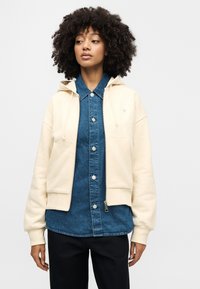 Εκρού φούτερ με κουκούλα και cropped σχεδιασμό, μαλακή υφή και μπροστινές τσέπες, φορεμένο πάνω από ένα μπλε τζιν πουκάμισο με κουμπιά πατιλέτα.