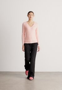 Gaiši rozā adīts džemperis ar V veida izgriezumu, kombinēts ar melnām biksēm ar svītrainu rakstu un rozā apaviem. Modelis soļo pretneitrālu fonu.