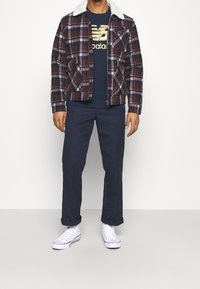 Ruta i flanelljacka med en vit shearlingkrage, marinblå t-shirt med guldlogotyp, marinblå byxor och vita sneakers med röda accenter.