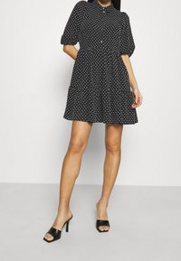Robe noire avec motifs de cœurs blancs, manches courtes bouffantes, col et taille froncée. Associée à des mules noires à talons hauts ouvertes au bout.