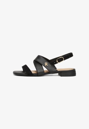Svarte sandaler med kryssende stropper, laget av lær og semsket skinn, med en lav blokkheel og en logo i gulltone på siden.
