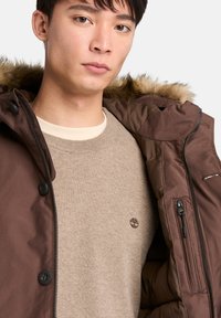 Brun isolerad jacka med huva i faux päls, med dragkedjestängning och knappdetaljer, som bärs över en beige crewneck-tröja.