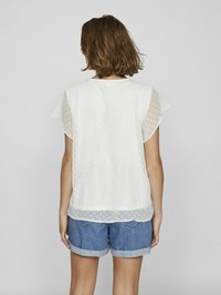 Blusa bianca semi-trasparente con motivo a pois, maniche corte a balze e orlo arrotondato, abbinata a pantaloni corti in denim.
