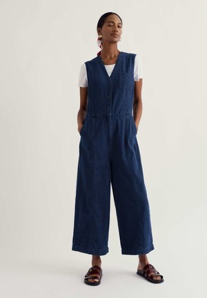 Žena oblečená v tmavomodrom rifľovom overale bez rukávov cez biele tričko, doplnená tmavohnedými sandálmi a zlatými kruhovými náušnicami.