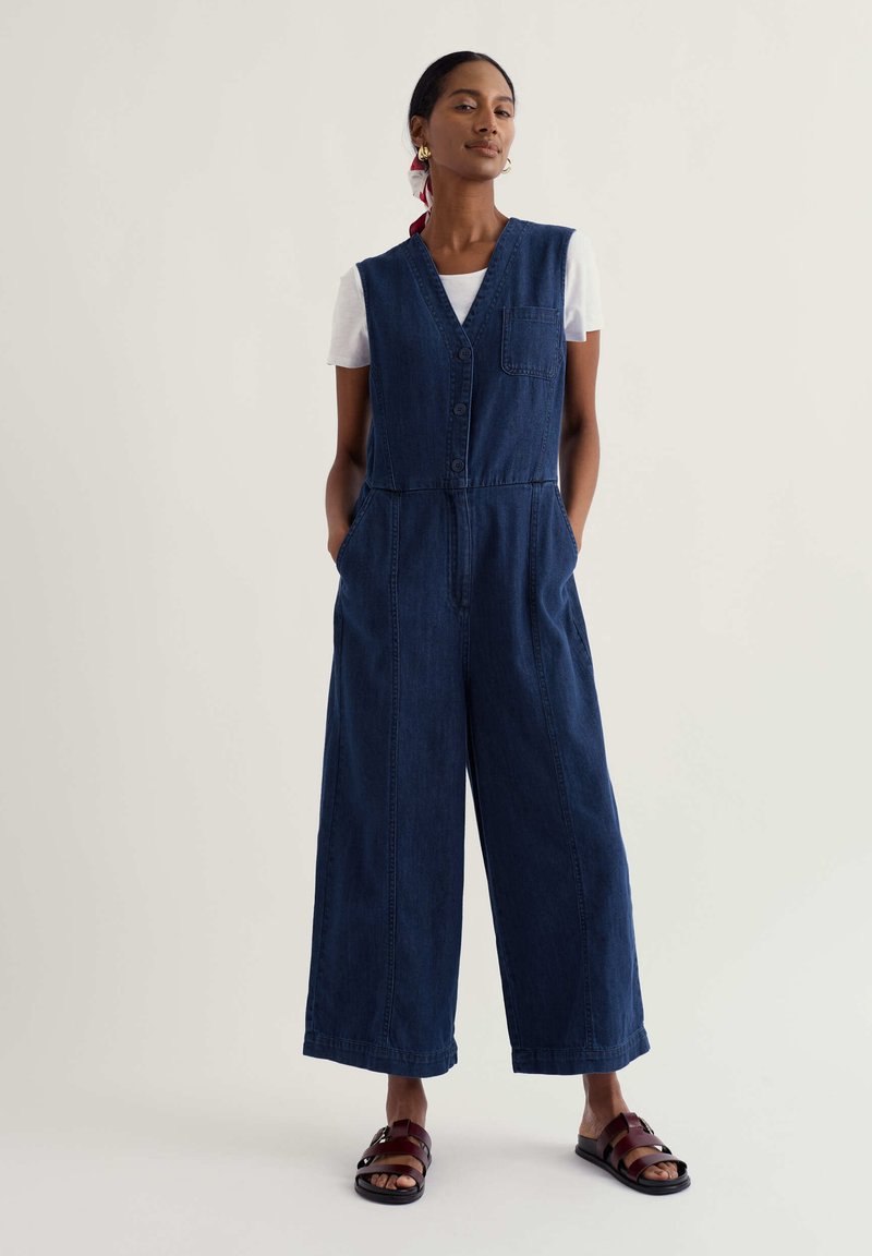 Frau trägt einen ärmellosen dunkelblauen Denim-Overall über einem weißen T-Shirt, kombiniert mit dunkelbraunen Sandalen und goldenen Creolen.