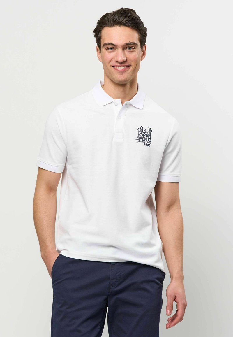 Jeune homme debout avec une main dans la poche, portant un polo blanc avec le logo "U.S. Open Polo Championship 2026" et un pantalon bleu marine.