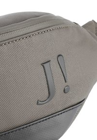 Šedá látková taška s texturovaným povrchem, s černým akcentním panelem na dně a výrazným šedým logem "J!" embosovaným na boku.