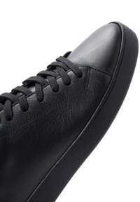 Zapatilla de cuero negro con una textura suave, puntera redondeada y cordones planos. Presenta una suela de goma texturizada y un discreto detalle de costura.