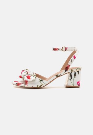 White heeled sandals with floral pattern featuring a bow on the toe, ankle strap with buckle, and a chunky block heel.