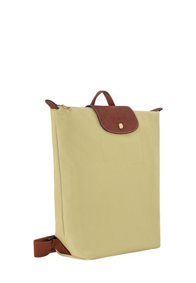 Hellgelber Rucksack aus weichem Material, mit einer braunen, strukturierten Klappe, einem goldenen Druckknopfverschluss und einem oberen Griff für einfaches Tragen.