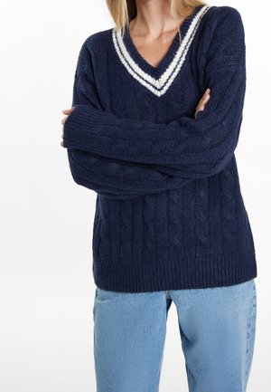 Personne portant un pull en maille torsadée bleu marine avec des rayures blanches sur le col en V et les bras croisés, associé à un jean bleu clair.