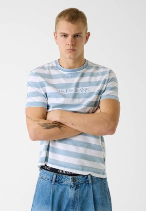 Jeune homme aux cheveux blonds courts portant un t-shirt Guess Jeans à rayures bleues et blanches et un jean bleu, bras croisés, expression neutre.