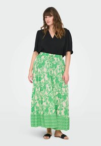 Top nero a scollo a V abbinato a una lunga gonna floreale verde e crema con orlo decorato. La modella indossa sandali neri.