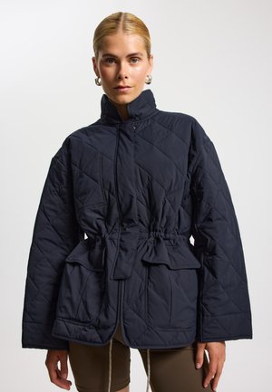 Granatowa pikowana kurtka z wysokim kołnierzem, taliowana w pasie i dużymi kieszeniami. Charakteryzuje się teksturowaną powierzchnią zewnętrzną oraz luźnym, strukturalnym krojem.