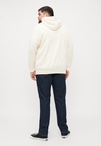 Kremfarget hettegenser med kengurulomme, ribbestrikkede mansjetter og kant, satt sammen med mørkeblå denimjeans og svarte sneakers. Utsikt fra baksiden.