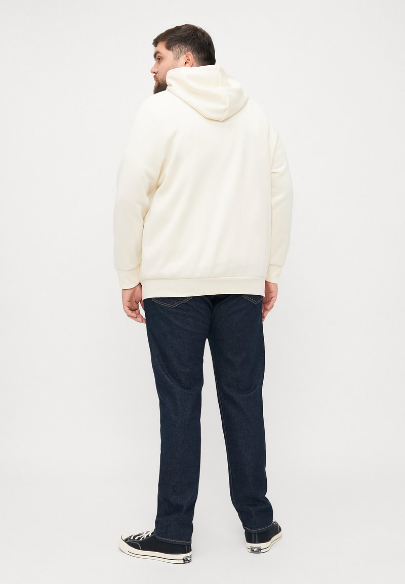 Sudadera crema con bolsillo tipo canguro, puños y dobladillo acanalados, combinada con jeans de mezclilla azul oscuro y zapatillas negras. Vista desde atrás.