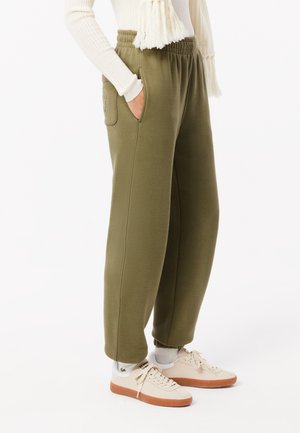 Olivgrüne Jogginghose mit elastischem Bund, Seitentaschen und strukturiertem Finish, kombiniert mit beigen Turnschuhen und weißen Socken.