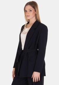 Blazer navy con cintura, silhouette strutturata, due tasche frontali e colletto a revers. Tessuto liscio con finitura opaca.