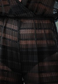 Zwarte sheer kledingstuk met verticale plooien en geometrische patronen, met afwisselend ondoorzichtige en transparante secties. Elastische tailleband zichtbaar.