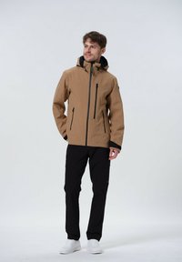 Tan waterdichte jack met een zwarte capuchon, twee voorzakken met rits en elastische manchetten. Gemaakt in combinatie met zwarte cargobroeken en witte sneakers.