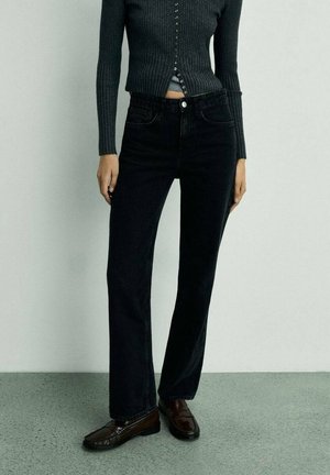 Person wearing fitted dark gray ribbed cardigan, high-waisted black straight-leg jeans, and dark brown loafers standing on gray floor.