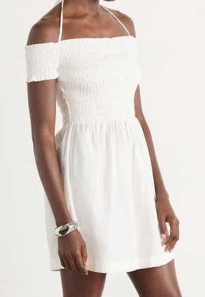 Femme portant une mini robe smockée blanche à épaules dénudées avec des bretelles halter et un bracelet argenté décalé au poignet droit.