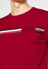 Červené bavlněné tričko s horizontálními pruhy v černé, bílé a červené barvě. Na tričku je potištěné logo s nápisem „TOMMY HILFIGER NEW YORK EST. 1985.“