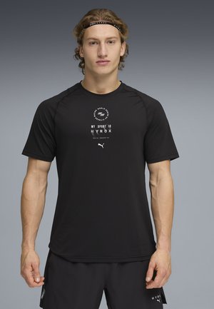 Atletisk mand med krøllet hår iført sort sportstøj og et "HYROX" pandebånd, stående foran en grå baggrund.