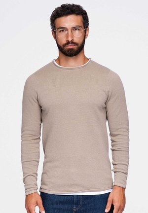 Homme avec barbe et lunettes porte une chemise beige texturée à manches longues et un jean bleu, debout devant un fond clair uni.
