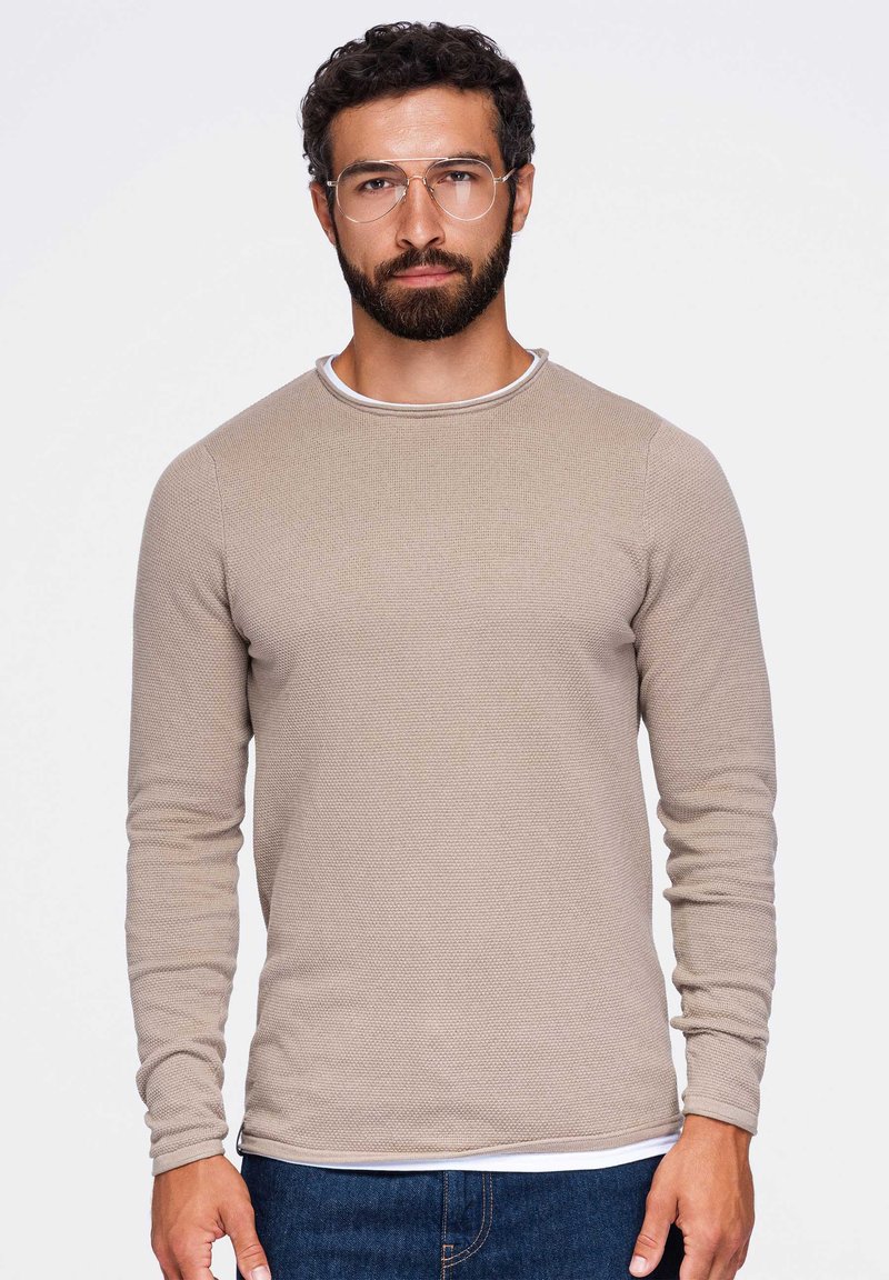 Homme avec barbe et lunettes porte une chemise beige texturée à manches longues et un jean bleu, debout devant un fond clair uni.
