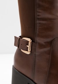 Bota alta de cuero marrón y tela con un detalle de hebilla dorada brillante y un tacón cuadrado negro, que presenta una textura suave y detalles de costura.