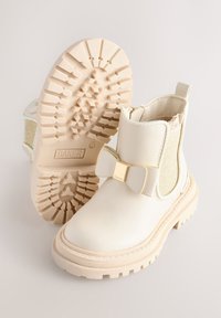 Bottines blanches en simili cuir avec un nœud doré en accent, un panneau latéral élastique texturé et une semelle en caoutchouc épaisse avec un motif de bande de roulement.