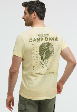 Mann trägt hellgelbes T-Shirt mit grünem Tiger und Text "Vietnam Camp David King of the Forest" auf dem Rücken.