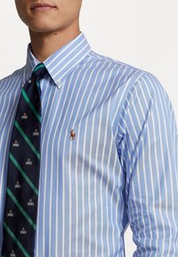 Camisa de cuadros azul claro y blanca con un pequeño logo bordado, combinada con una corbata azul marino que presenta un patrón diagonal en verde y azul.