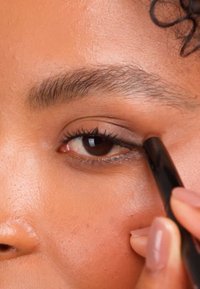 Zbliżenie na oko osoby z brązową tęczówką, nakładającej czarną kredkę do oczu. Skóra ma gładką fakturę i ciepły odcień.