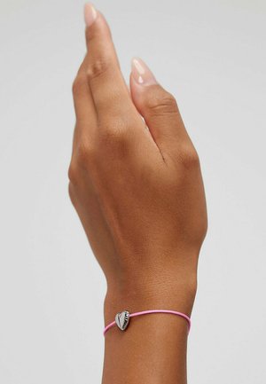 Bracelet élastique rose avec un charme en forme de cœur en argent, présentant une surface lisse et un design minimaliste, porté sur une main.