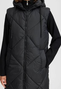 Schwarze gepolsterte Weste mit einem gesteppten Rautenmuster, Kapuze und Frontreißverschluss. Verfügt über zwei Seiten Taschen und verstellbare Kordelzüge an der Kapuze.
