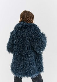 Dlhý, textúrovaný kabát s huňatým, tmavomodrým materiálom pripomínajúcim kožušinu. Má široký golier a voľný strih, končiaci v oblasti bokov.