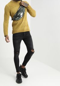 Żółty dziergany sweter, czarne podarte obcisłe jeansy, czarne trampki oraz zielona moro nerka z białym logo Nike.