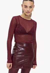 Bordeaux lange mouw mesh top met zilveren studs, gelaagd over een bijpassende bralette, gecombineerd met een hoge taille, glanzende bordeaux leren minirok.