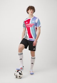 Maillot de sport bleu, rouge et blanc avec des motifs abstraits ; shorts noirs ; chaussettes blanches avec des rayures noires ; ballon de football noir et blanc.