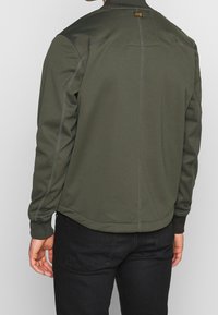 Olivgrön jacka med en slät yta, ribbade manschetter och hög krage. Har kontrastsyddning och en logotags bakpå.