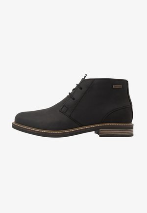 Schwarze Lederknöchelstiefel mit runder Spitze, zwei Schnürösen, beigem Stich und einer strukturierten, dunkelbraunen Sohle. Barbour-Logo auf der Außenseite.