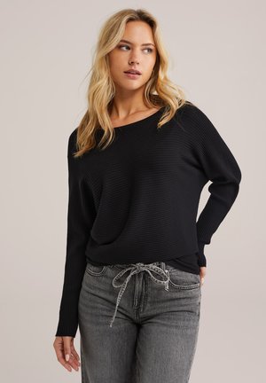 Schwarzes, geripptes Langarmoberteil mit einem runden Ausschnitt, kombiniert mit grauen, hochtaillierten Jeans, die einen gebundenen Kordelzug zum Schließen haben.