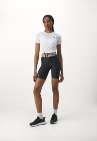 Hvid kortærmet top med knude-detail, parret med højtaljede sorte cykelshorts med iøjnefaldende hvid tekst på waistbandet. Sorte træningssko.