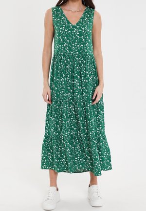 Frau trägt grünes ärmelloses Midi-Kleid mit V-Ausschnitt und weißem Blumenmuster sowie weißen Sneakers, steht vor einem schlichten weißen Hintergrund.