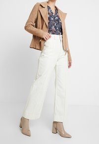 Braune Wildleder-Bikerjacke, florales Hemd, weiße weit geschnittene Hosen und beigefarbene Stiefeletten mit Absatz. Strukturiertes Material und klare Linien.