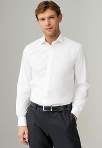 Chemise blanche à manches longues avec un col classique, en coton, texture lisse, portée avec un pantalon foncé et une ceinture noire.