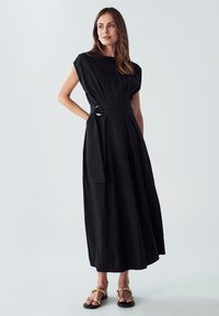 Črna, kratka obleka maksi dolžine s pripetim pasom in detajlom pasu. Narejena iz teksturirane tkanine z prosto obliko.