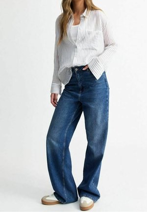Persoon die wijde blauwe jeans draagt, witte sneakers, een wit hemdje en een los wit overhemd met dunne zwarte strepen.